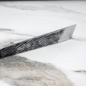The V2 Essential Skiver, a single-edged knife with a patterned blade, rests on a white marble surface.