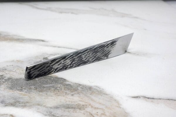 The V2 Essential Skiver, a single-edged knife with a patterned blade, rests on a white marble surface.