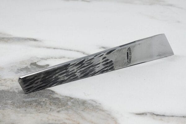 Close-up of The V2 Essential Skiver, a stainless steel hair comb with a textured pattern, resting on a light marble surface.