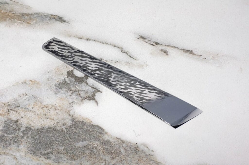 The Essential Skiver, a polished rectangular metallic tool featuring a textured pattern, rests on a marbled surface.
