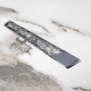 The V2 Essential Skiver, a decorative metal strip with a geometric pattern, rests on a marbled white and gray surface.