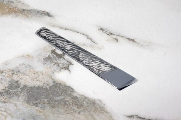 The V2 Essential Skiver, a decorative metal strip with a geometric pattern, rests on a marbled white and gray surface.