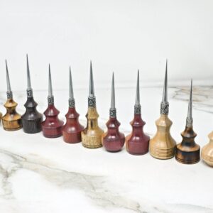 A row of eleven Premium Handmade to Order Scratch and Poking Awls with wooden handles in various shades, featuring metal tips, placed on a marble surface next to a ruler and a metal tool.