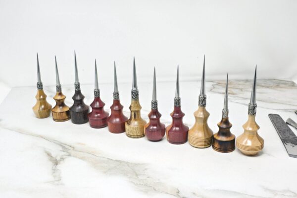 A row of eleven Premium Handmade to Order Scratch and Poking Awls with wooden handles in various shades, featuring metal tips, placed on a marble surface next to a ruler and a metal tool.