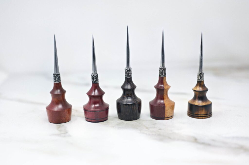 Five Premium Handmade to Order Scratch and Poking Awls with wood handles and metal tips stand upright on a white marble surface, each featuring different shades and grain patterns of wood.