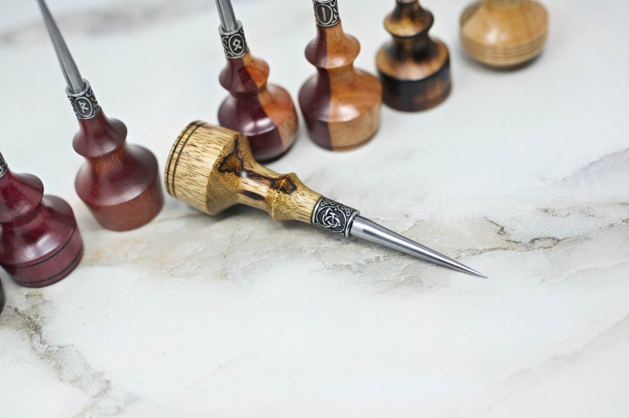 Various wooden-handled Premium Handmade to Order Scratch and Poking Awls displayed in a line on a light marbled surface, with one poking awl positioned horizontally in the center foreground.