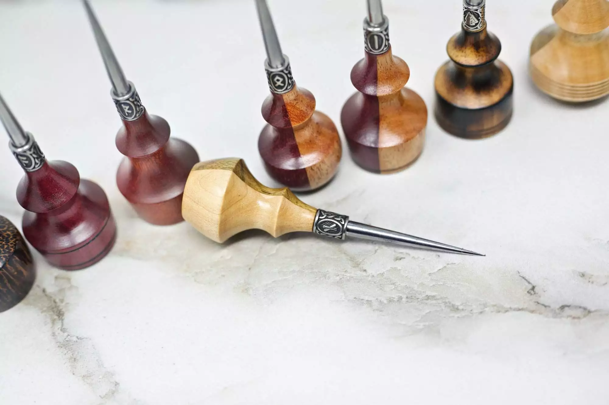 A collection of Premium Handmade to Order Scratch and Poking Awls with wooden handles and metal tips, displayed in varying shades and grain patterns, arranged on a light marble surface.