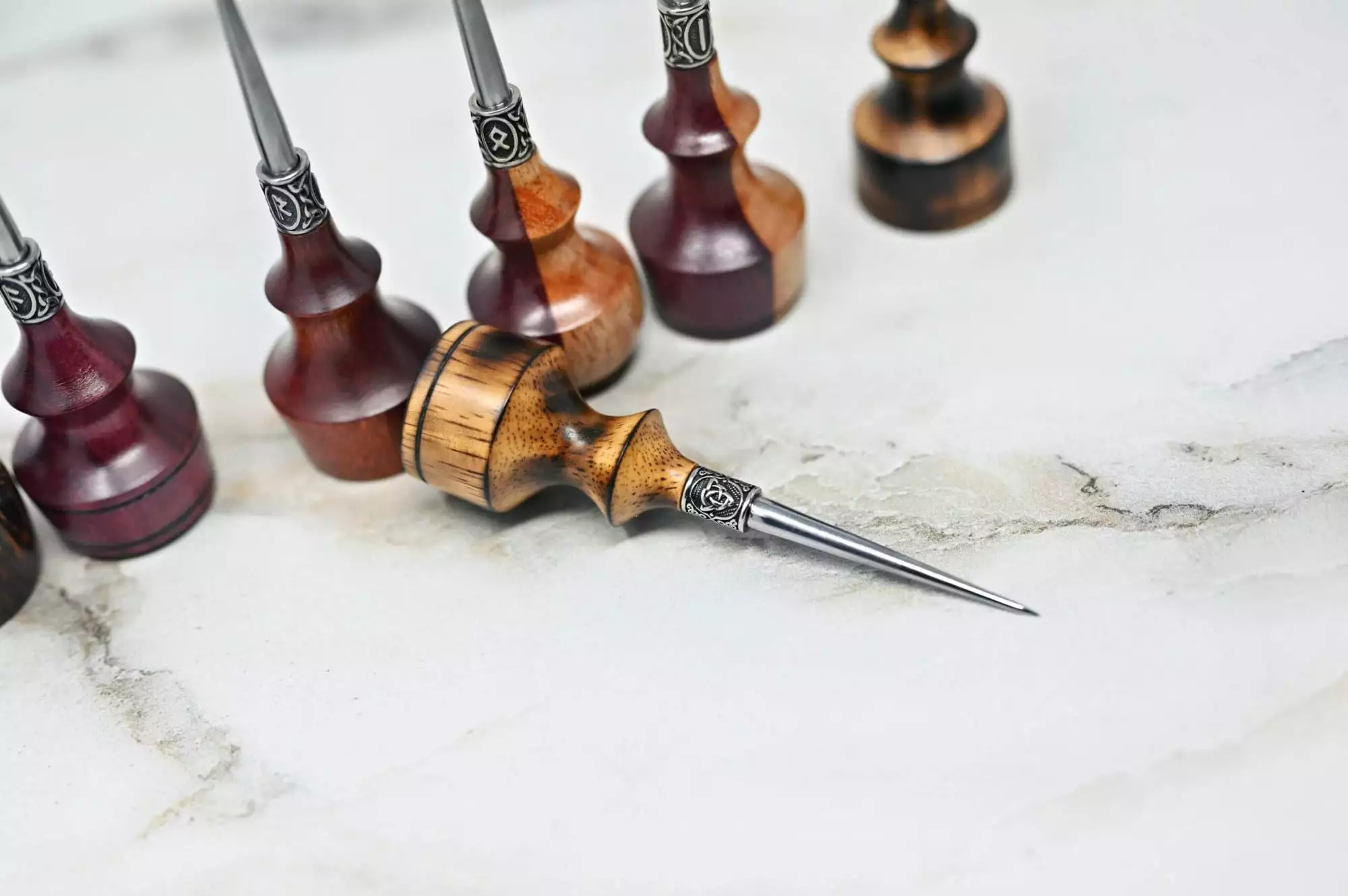 A row of Premium Handmade to Order Scratch and Poking Awls with wood handles and various finishes, placed on a light-colored marble surface.