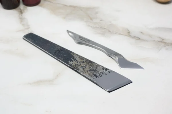 Two straight razors are laid out on a marble surface: a rectangular Fang with a worn blade and an Essential Skiver, smoother modern one with a distinct curve.