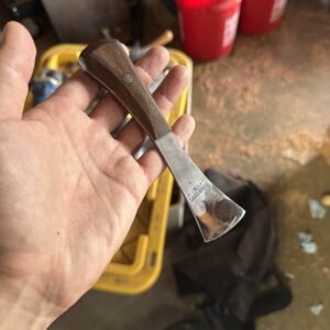 A hand holding a chisel with a wooden handle in a workshop setting. Red buckets and various tools, including the Labor Deal 7 Piece Special set, are visible in the background.