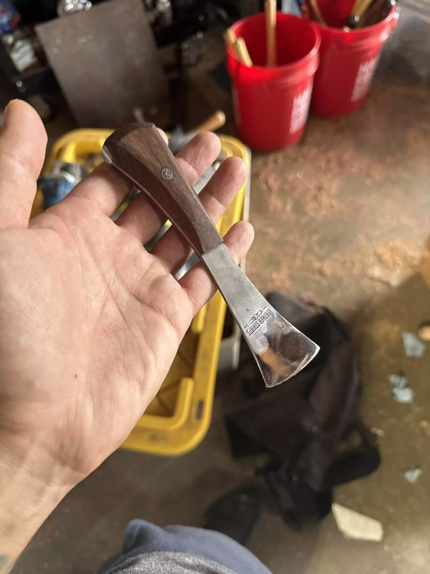 A hand holding a chisel with a wooden handle in a workshop setting. Red buckets and various tools, including the Labor Deal 7 Piece Special set, are visible in the background.