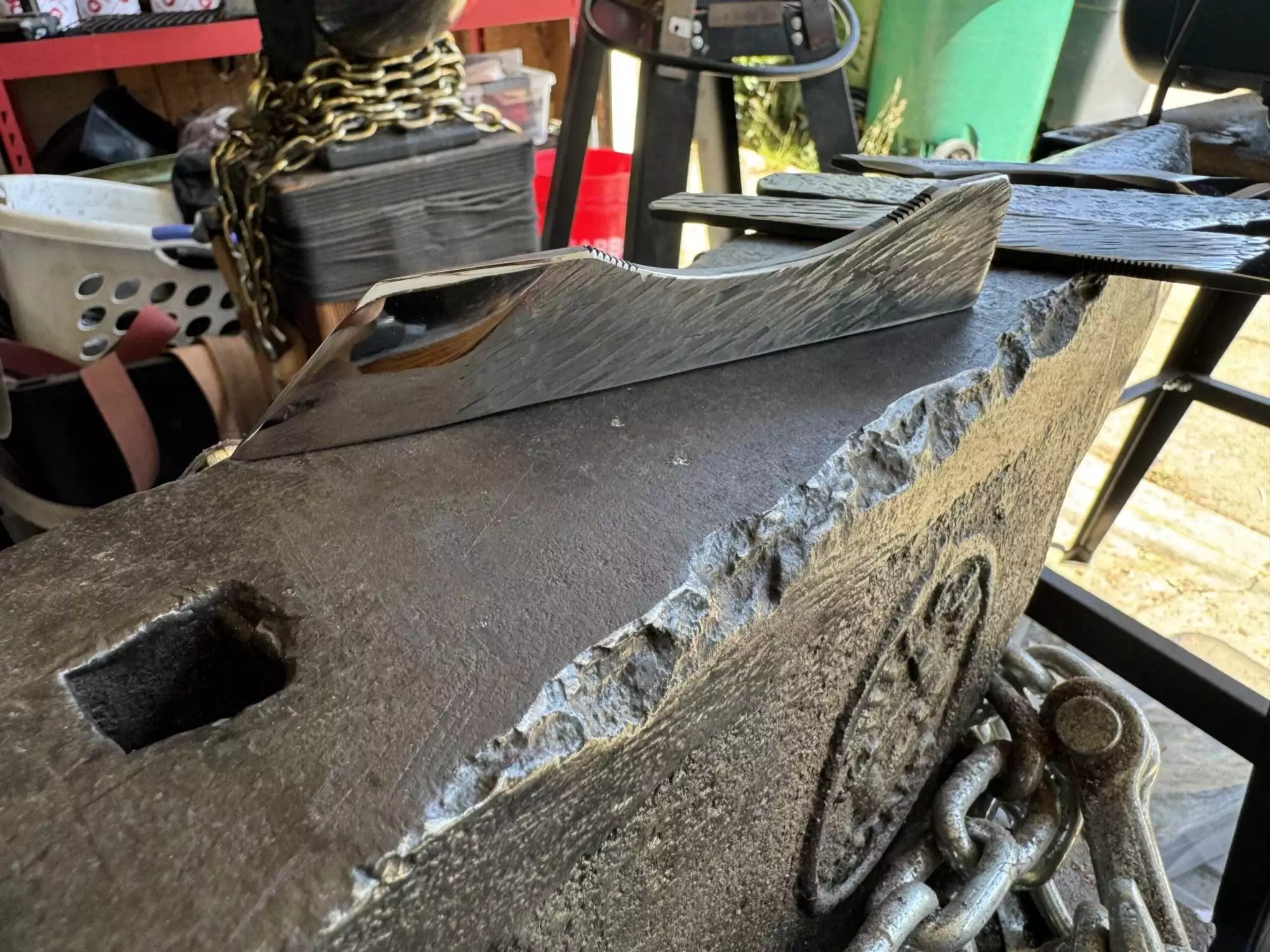 In a workshop, a metal plane from the Labor Deal - 7 Piece Special rests on a large, rough-edged anvil. Chains, tools, and storage items can be seen in the background.