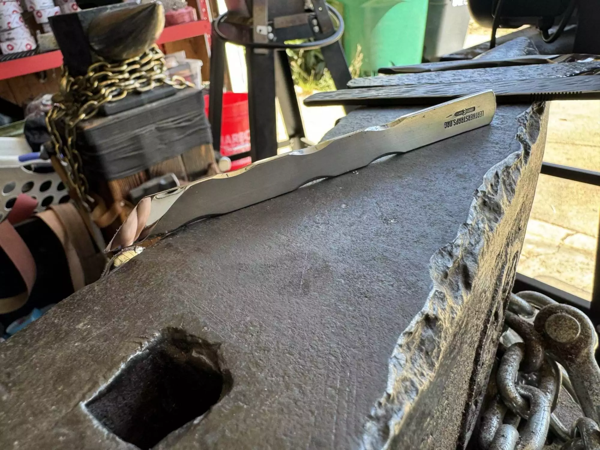 Close-up of a serrated blade from the Labor Deal - 7 Piece Special resting on a rough-textured anvil in a workshop setting, surrounded by various tools and chains.