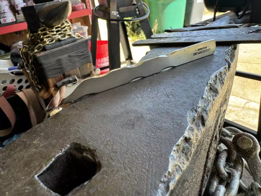 A forge featuring the Labor Deal 7 Piece Special, showcasing an anvil with various metalworking tools, including a flat bar resting on the anvil and a hammer nearby. Chains and equipment are visible in the background, evoking the essence of a craftsman's dedicated labor deal.