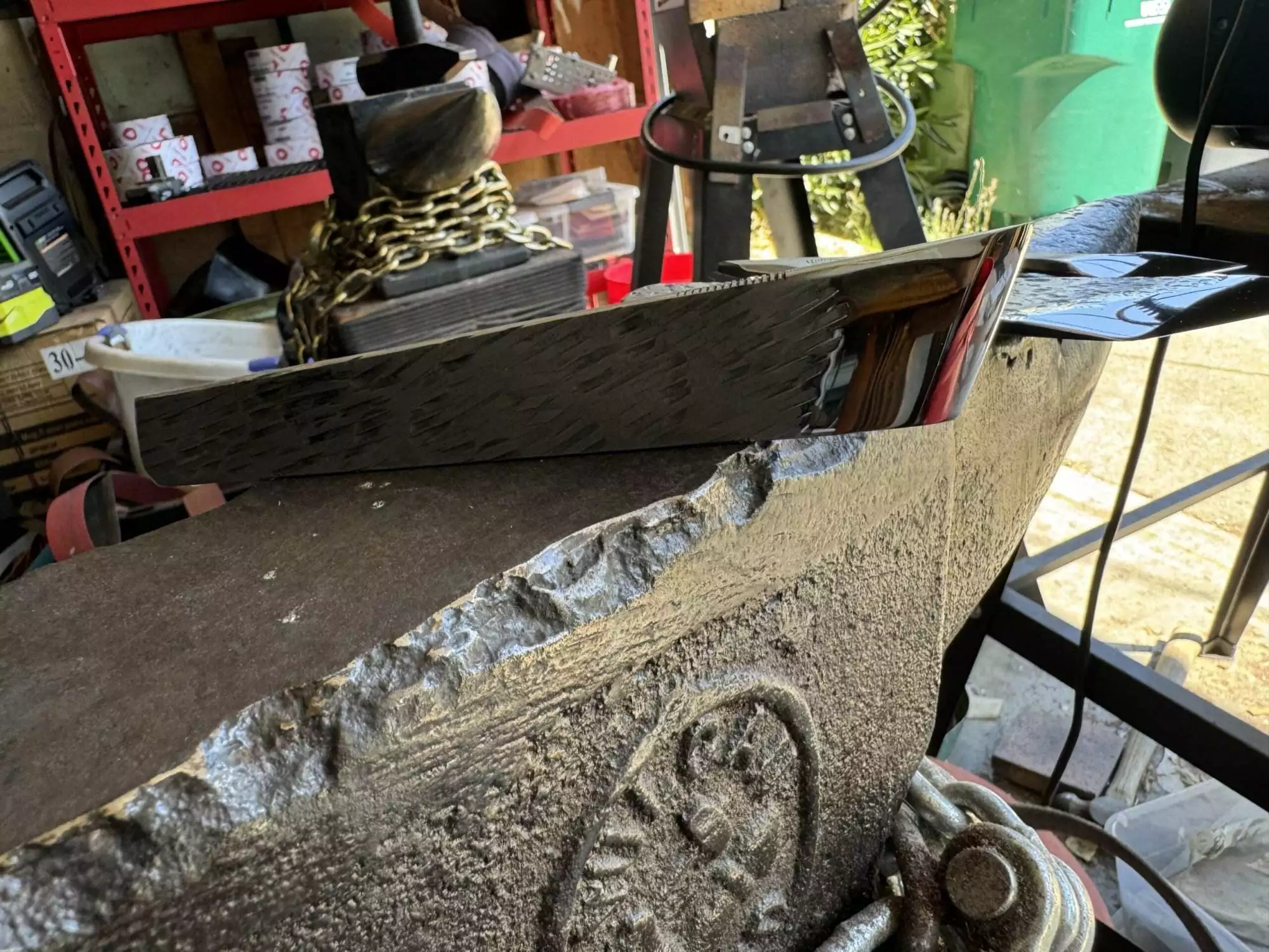 A polished metal blade from the Labor Deal - 7 Piece Special sits atop a heavily used anvil in a cluttered workshop, with chains and various tools visible in the background.