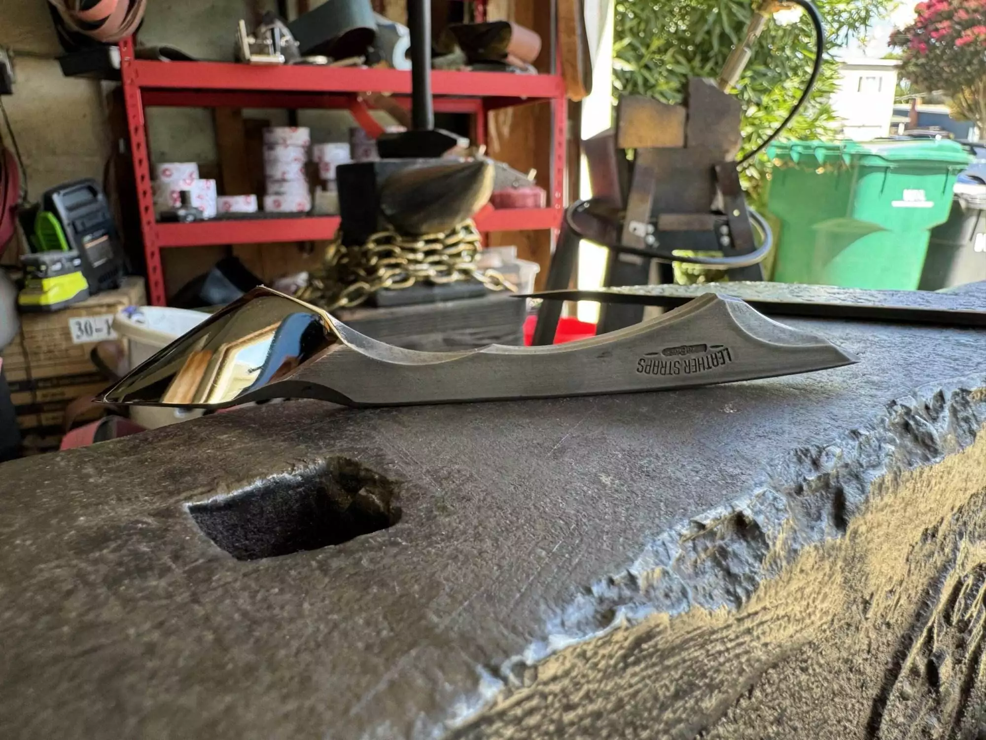 A knife handle from the "Labor Deal - 7 Piece Special" set, engraved with "TBT Forge + Press," rests on an anvil in a cluttered workshop surrounded by various tools and equipment.