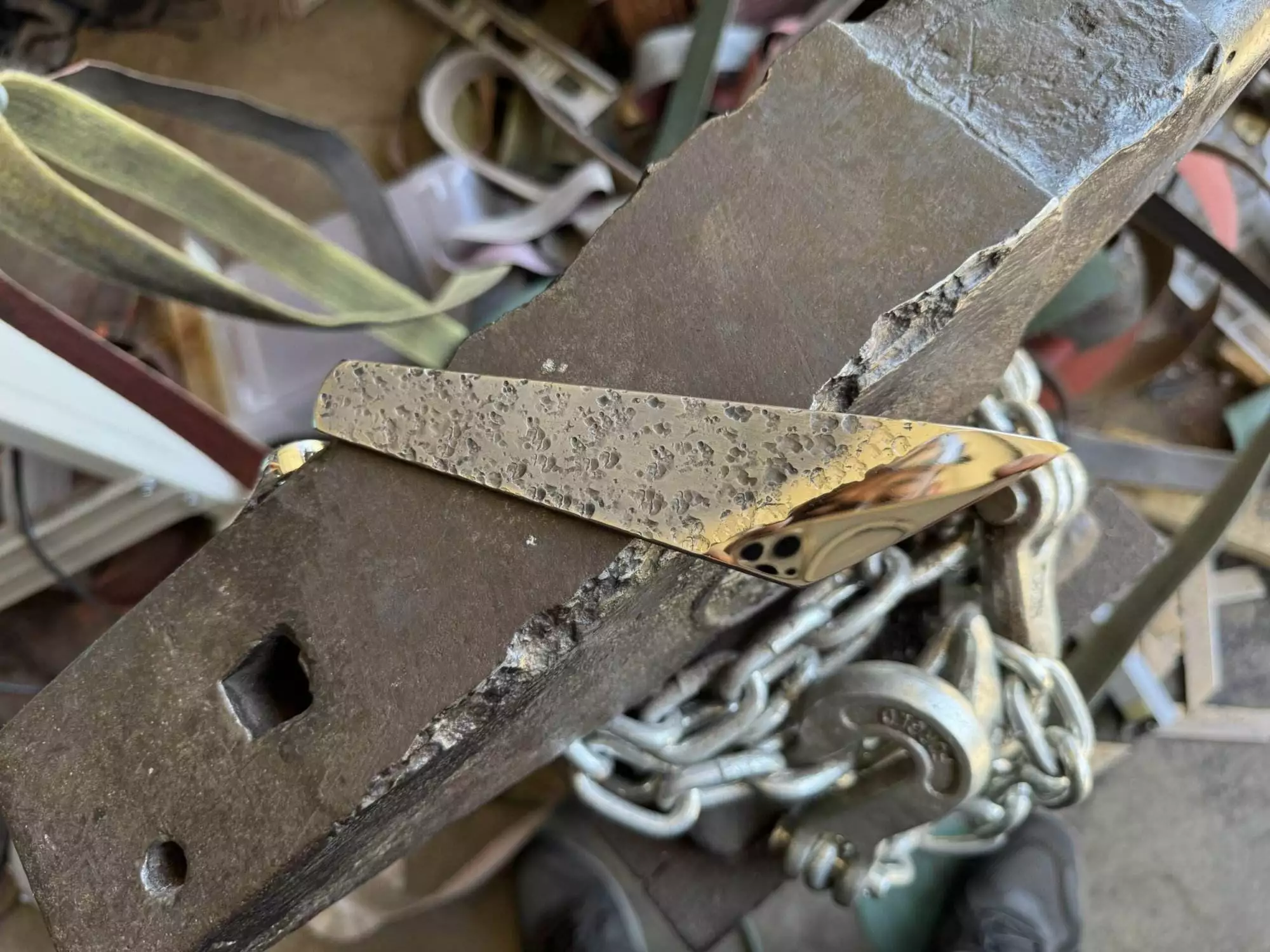 A piece from the "Labor Deal - 7 Piece Special" collection, featuring a shiny, gold-colored metal bar with a textured surface, lies diagonally on a heavily-worn, rusted metal block near a chain on a workshop table.