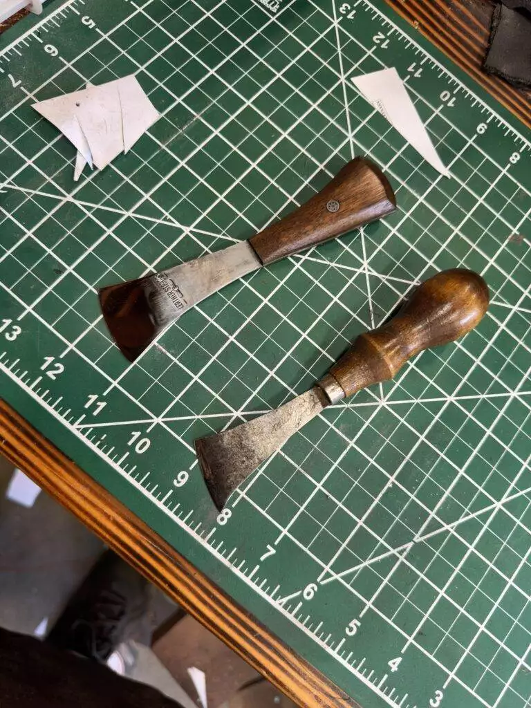 Two wood carving chisels with wooden handles are placed on a green cutting mat with a grid pattern. Scattered scraps of paper and part of a wooden table are also visible, reminiscent of tools from our Labor Deal 7 Piece Special set.