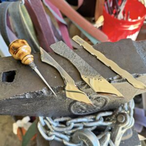 A set of metal pieces and a wooden-handled awl rest on an anvil, with a background of various tools and materials in a workshop. Don't miss our Exclusive Friday the 13th Sale Bundle, perfect for all your crafting needs.