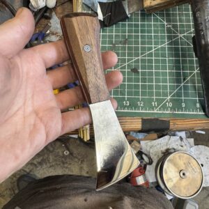 A person holds the "Signature" Leather Knife accompanied by a handmade awl. The background features a cutting mat and various tools.