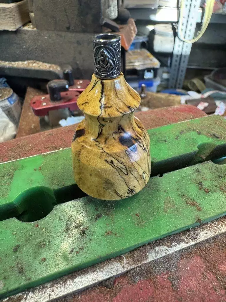 A wooden bottle with a decorative metal cap sits as a centerpiece on a green workbench in a cluttered workshop, surrounded by tools like the "Signature" Leather Knife and Spalted Tamarind Awl.