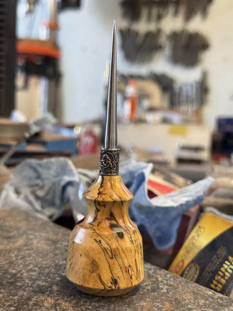 The "Signature" Spalted Tamarind Awl, featuring a polished, pointed metal tip and a smooth, patterned wooden handle, is prominently displayed on a workbench within the workshop setting.