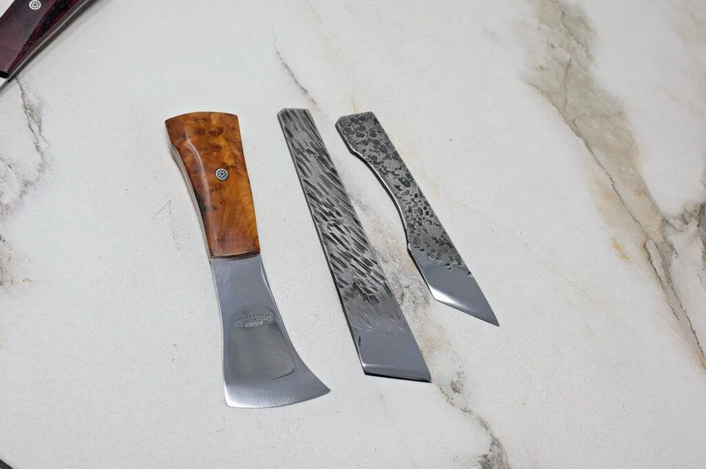 Three unique metal kitchen tools featuring wooden and textured handles are beautifully displayed on a light marble surface, forming "The Halloween 3 Piece Special" for your spooky season cooking adventures.