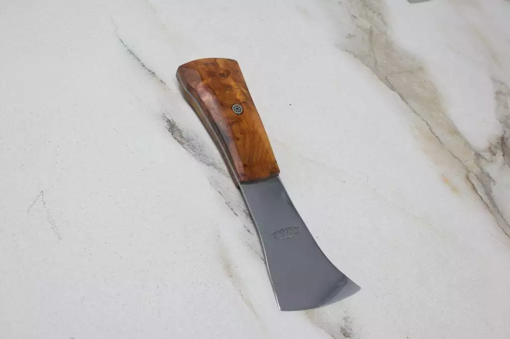 The 'Signature' Leather Knife, featuring a curved design with a polished wooden handle and a metal blade, rests on a marble surface.