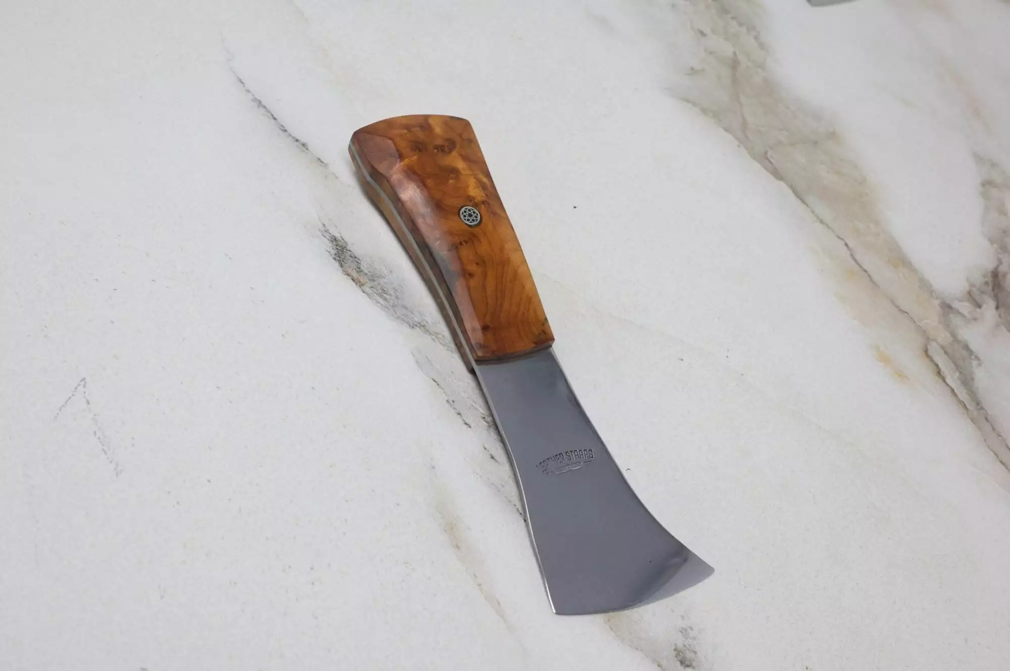 The 'Signature' Leather Knife, featuring a curved design with a polished wooden handle and a metal blade, rests on a marble surface.