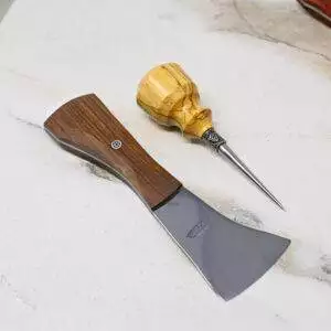 A small axe featuring a wooden handle and a woodworking awl rest on the white marble surface, completing The Halloween 3 Piece Special arrangement.