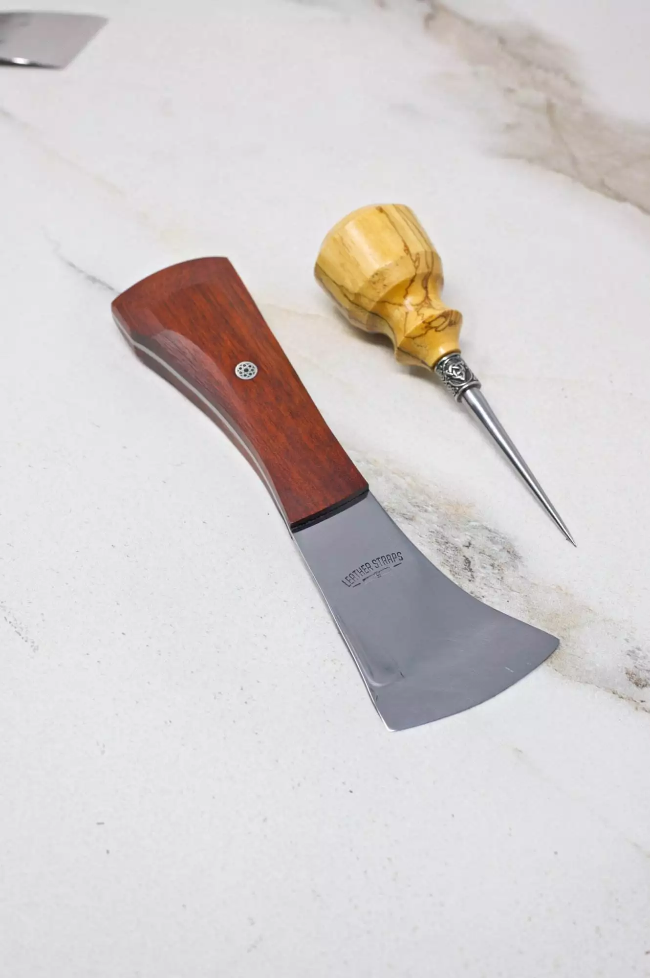 A petite axe with a wooden handle lies next to an awl, also equipped with a wooden handle, on a light marble surface, creating the ideal setup for The Halloween 3 Piece Special.