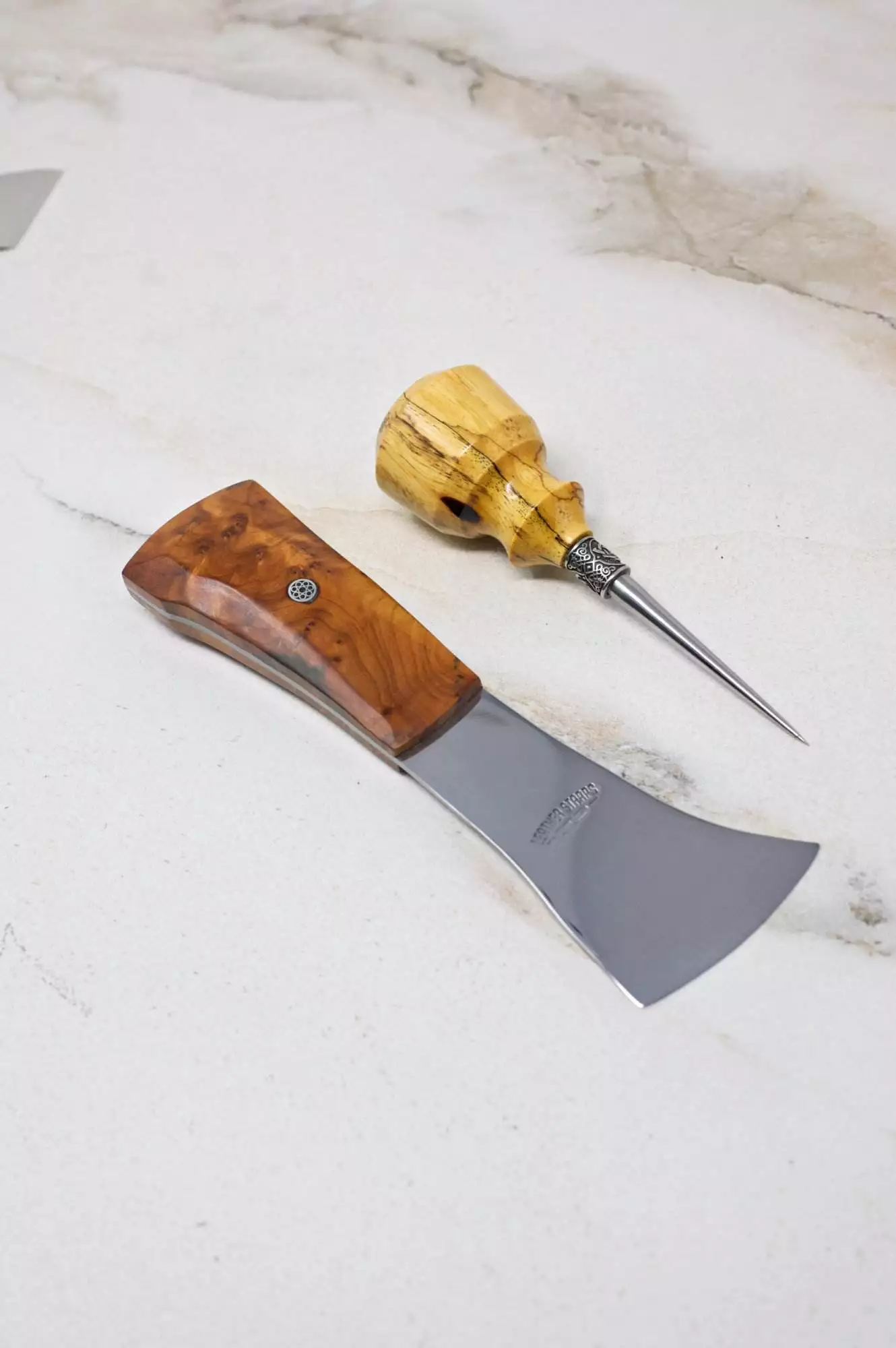 A woodworking chisel with a wooden handle and a carving tool featuring a swirl-patterned grip are displayed on a marble surface, part of The Halloween 3 Piece Special.