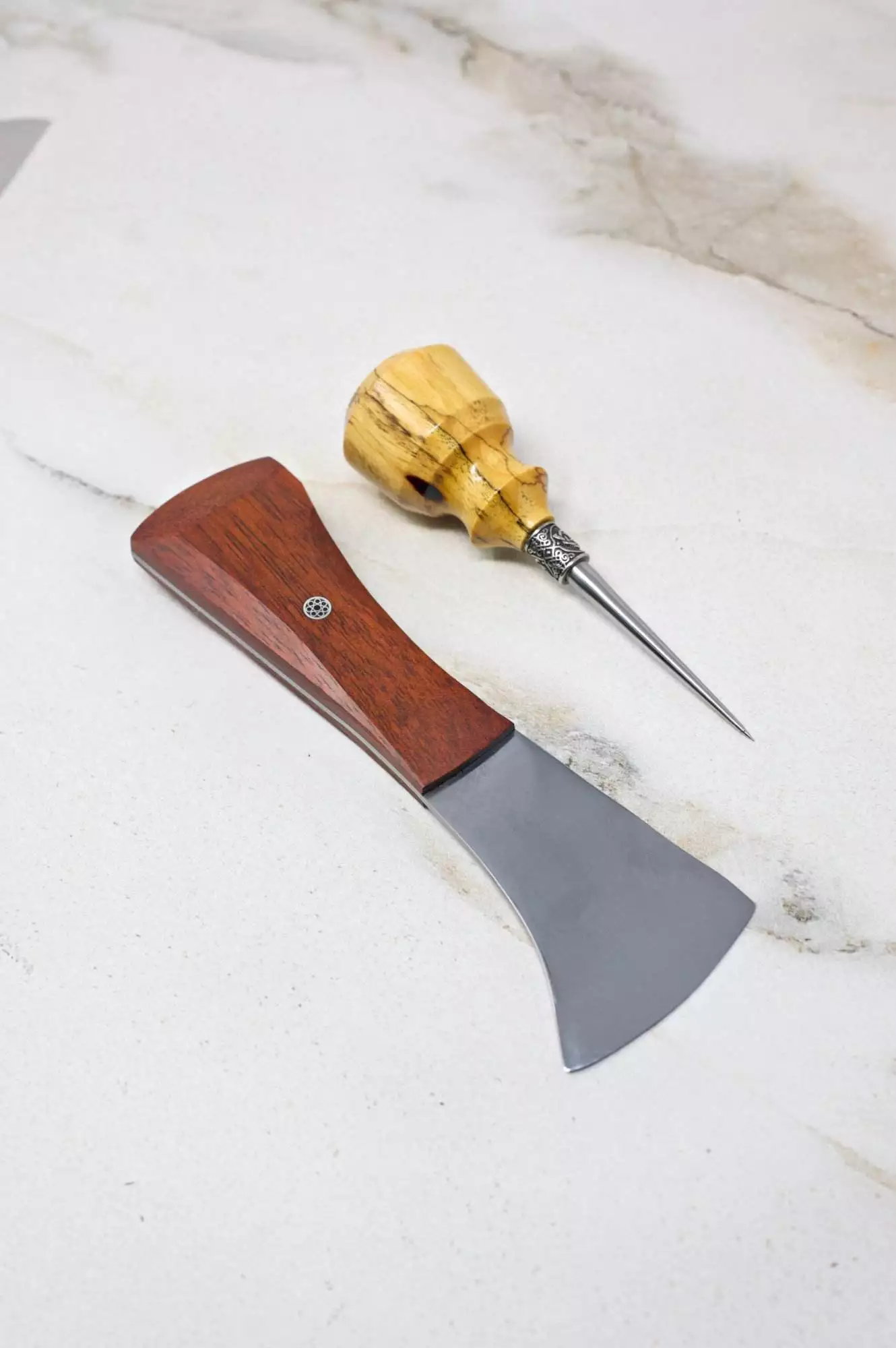 On a marble surface, as part of The Halloween 3 Piece Special, a small axe and a wood-handled awl are arranged.