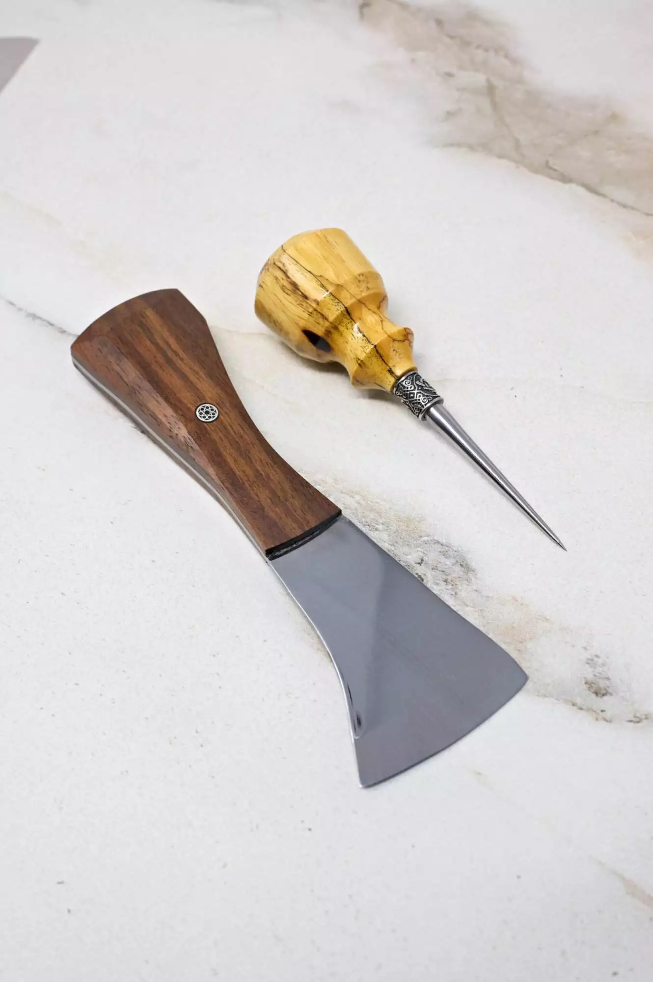 On a light marble surface, a small axe featuring a wooden handle is placed alongside an awl with a matching wooden grip. This pair, ideal for creating eerie Halloween decorations, might be just what your collection lacks—envision it as the missing piece to complete The Halloween 3 Piece Special.