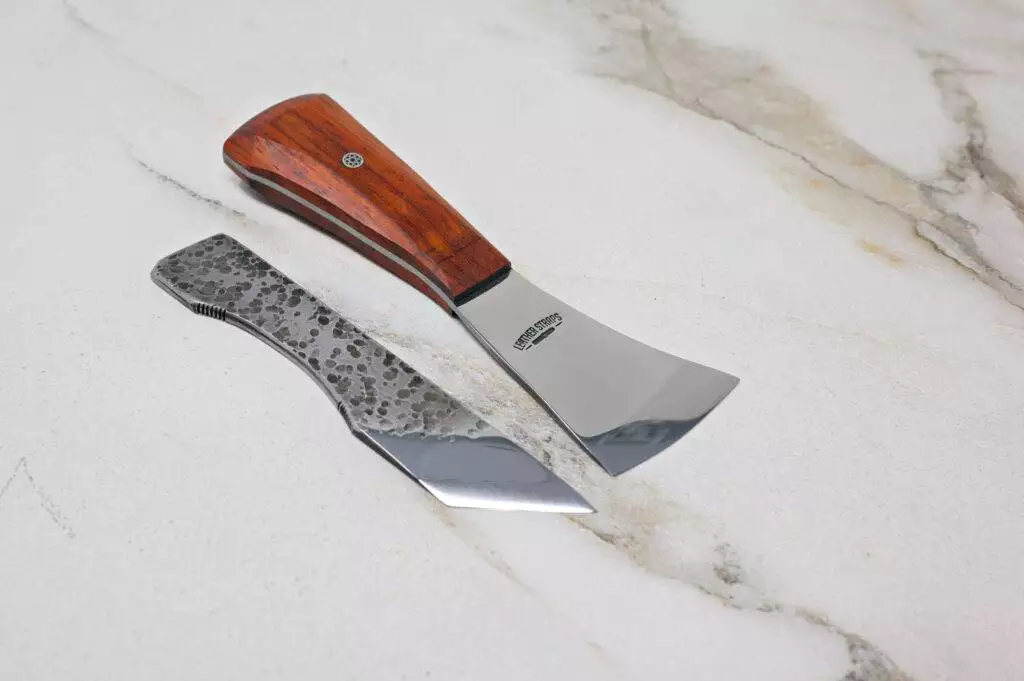 On a marble surface, two knives evoke the spirit of Halloween: one features a wooden handle with a wide blade, while the other has a speckled metal handle and narrow blade. They are part of The Halloween 3 Piece Special set.