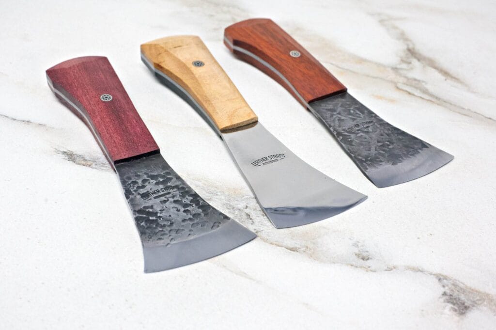 Three American Leather Knives, each featuring unique wooden handles and elegantly polished metal blades, are displayed on a marble surface.