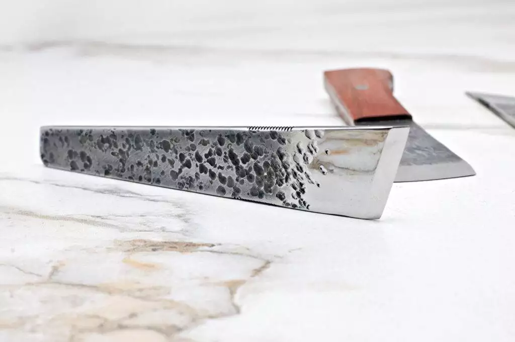 Close-up of The "Signature" American Leather Knife on a marble surface, showcasing its textured metallic blade and wooden handle. Another knife is partially visible in the background.