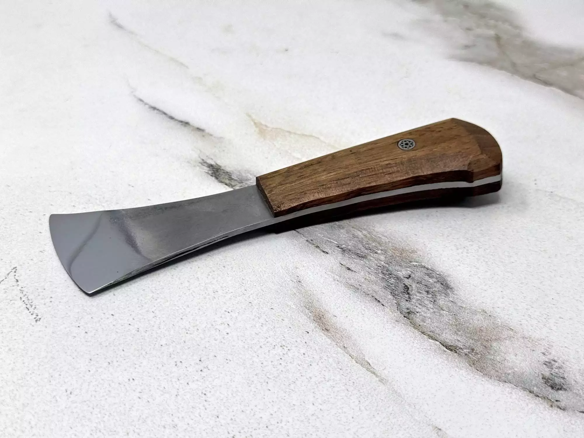 A compact stainless steel putty knife with a wooden handle, echoing the elegance of The "Charlie" Leather Knife + Handmade Awl, rests on a marbled surface.