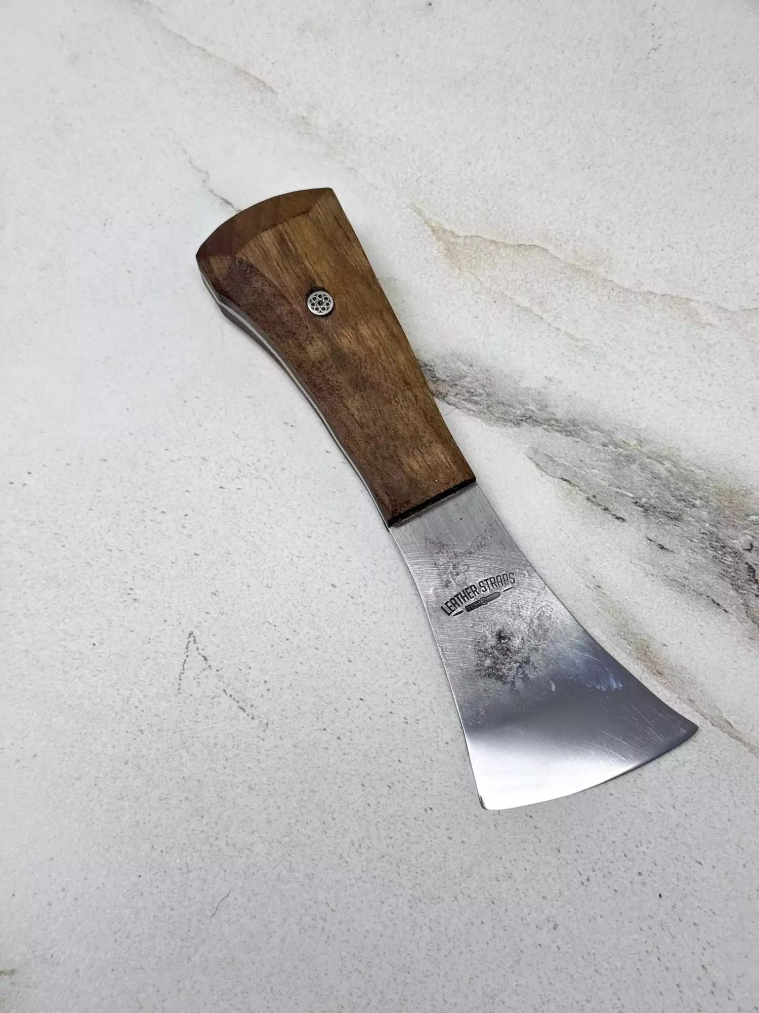 The "Charlie" Leather Knife, featuring a wooden handle and a short, curved blade, rests elegantly on a marble surface.