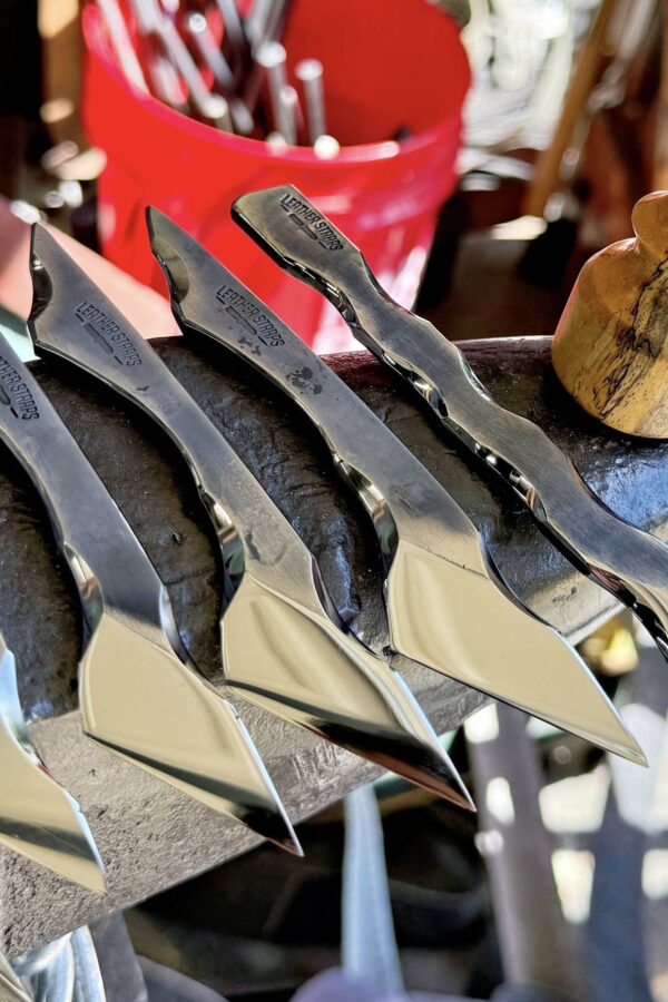 Four polished metal blades from "The Signature" Full Set rest on a black surface, with a blurred red bucket in the background.