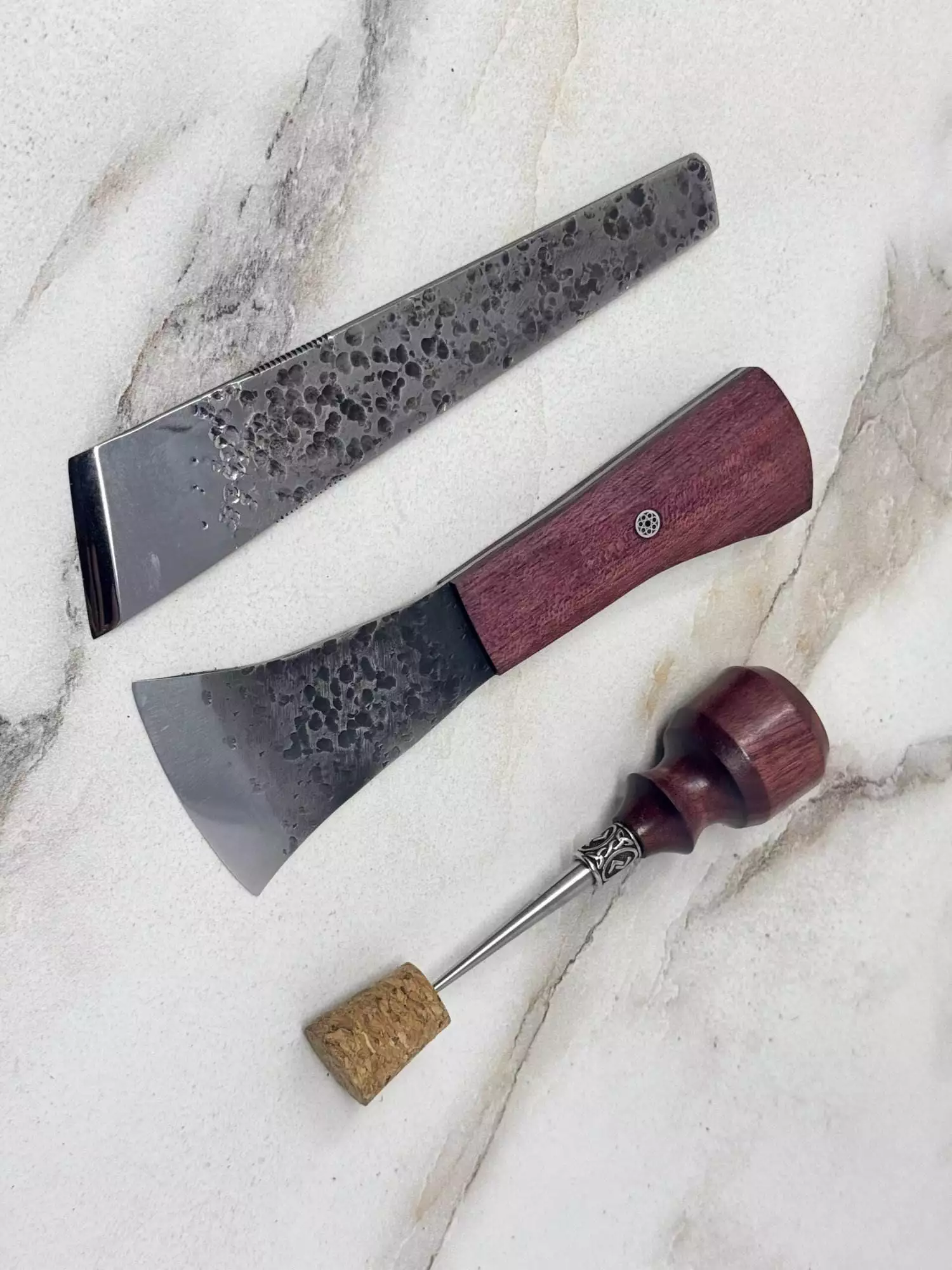 A forged custom three-piece metal and wood carving tool set for left-handed users, featuring a cork with a decorative pin, elegantly rests on the marble surface.