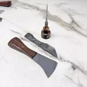 A set of woodworking tools on a marble surface includes the Black Walnut Signature Knife, Burnt Ambrosia Maple Awl, and two chisel blades with wooden handles.