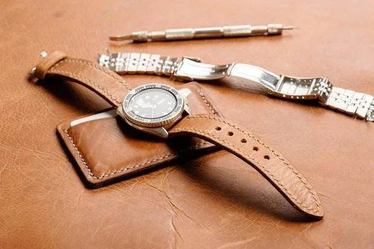 A wristwatch with a brown leather strap rests on a wallet, accompanied by a metal watch strap and a watch tool. All are placed on an expertly crafted leather surface, reminiscent of fine leathercraft.