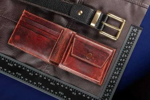 A brown leather wallet with clean corner cuts lies open on a dark surface next to a black belt with a gold buckle. A ruler, essential for precise leathercraft, is placed beside them.