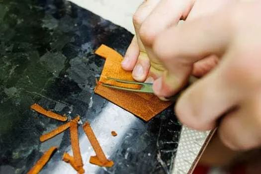 A person skillfully uses tweezers in leathercraft, handling thin strips of brown material on a dark surface to achieve clean corner cuts.