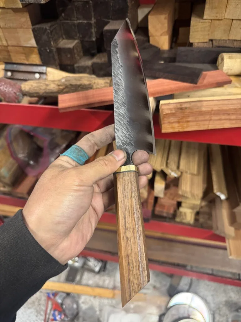A hand grips "2025 Valentine's Day - The Essentials Special," a chef's knife with a wooden handle, poised before shelves of diverse wood pieces, ready for crafting culinary creations.