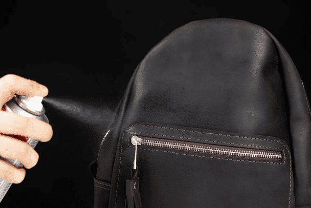 A hand skillfully sprays a can onto the black leather backpack, enhancing its water resistance. The zipper pocket gleams, a testament to fine leathercraft.