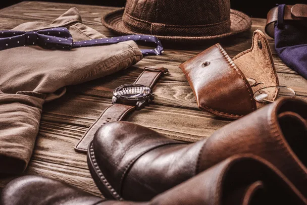 Men's fashion accessories, crafted with exquisite leatherworking, rest stylishly on a wooden surface, featuring a hat, watch, leather shoes, belt, eyeglasses case, bow tie, and trousers.
