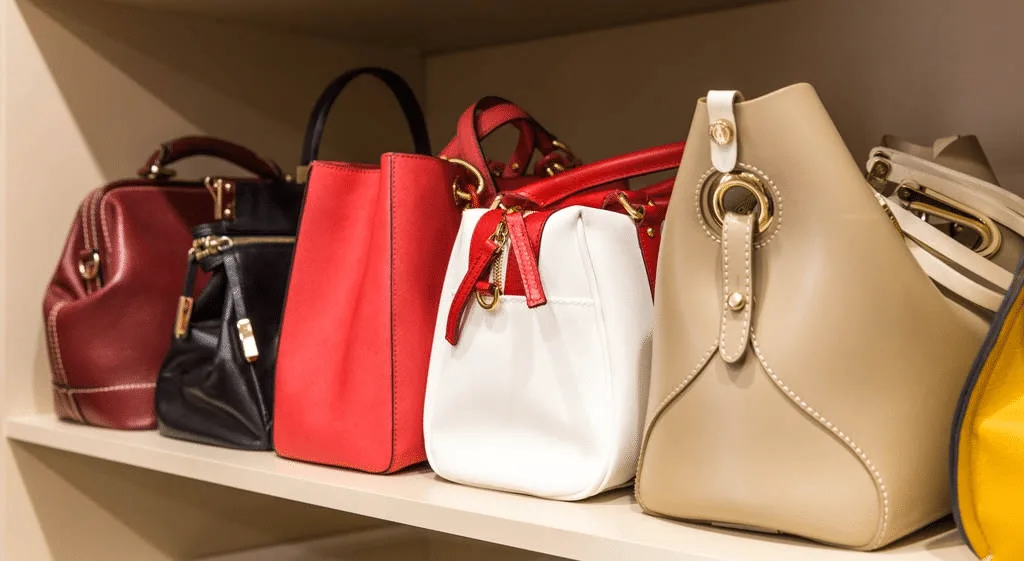 A shelf displaying various handbags in red, black, white, and beige shades showcases exquisite leathercraft, each piece neatly arranged.