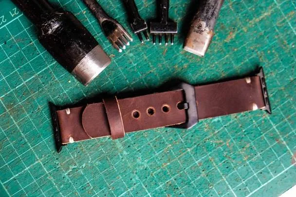 Brown leather watch strap on a green cutting mat with various leatherworking tools above it, showcasing the art of leathercraft and precision edge finishing.